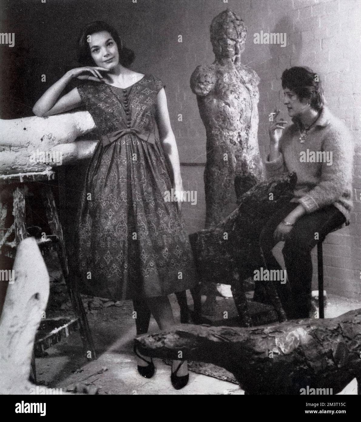 Ein Model, das ein Baumwollkleid von Sambo trägt, besucht Elisabeth Frink, die Bildhauerin in ihrem Atelier. Frink macht keine Abstriche daran, dass sie in einem Tatler-Modeshow gezeigt wird und im Hintergrund auf einer Zigarette sitzt. Stockfoto