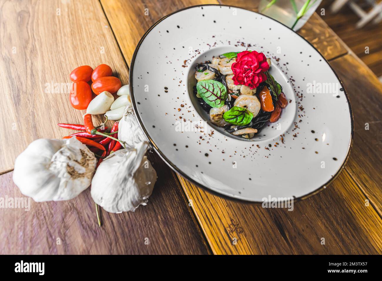 Garnelen-Gericht. Schwarze Tagliolini-Pasta mit gebratenen Garnelen, dekoriert mit essbaren Blumen. Weiße Platte auf Holztisch. Hochwertiges Foto Stockfoto