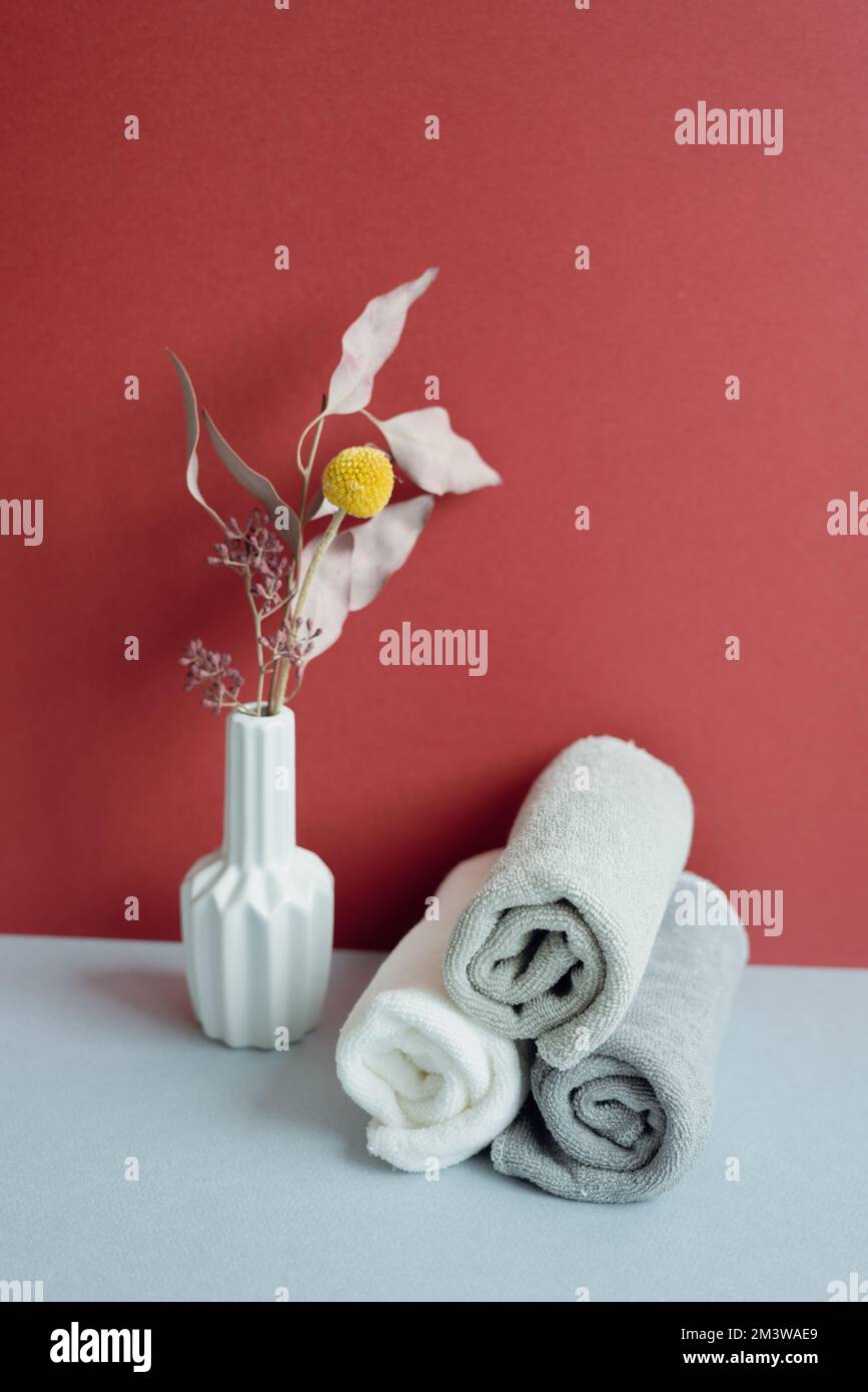 Ein Stapel gerollter Duschhandtücher aus Baumwolle und eine Vase trockener Blumen. Spa-Konzept für Badezimmer. Roter und grauer Hintergrund Stockfoto