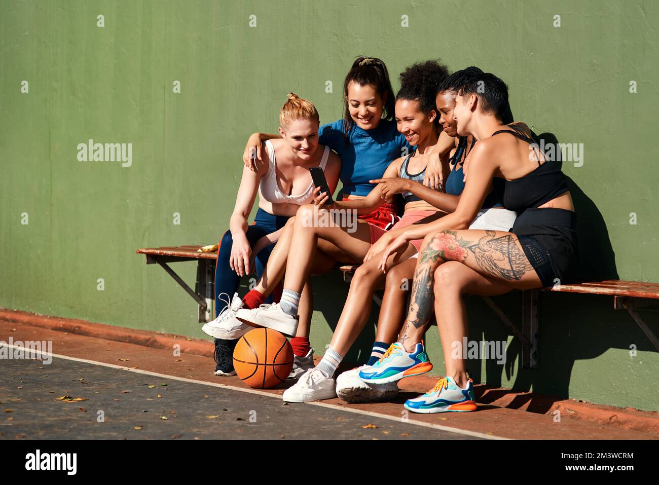 Lächeln. Eine vielfältige Gruppe von Sportlerinnen, die zusammen sitzen, nachdem sie Basketball gespielt und ein Handy angesehen haben. Stockfoto