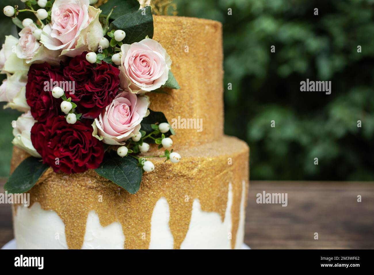 Zweistufige Hochzeitstorte in Weiß und Gold mit roten und pinkfarbenen Rosen und Zruskusblättern Stockfoto