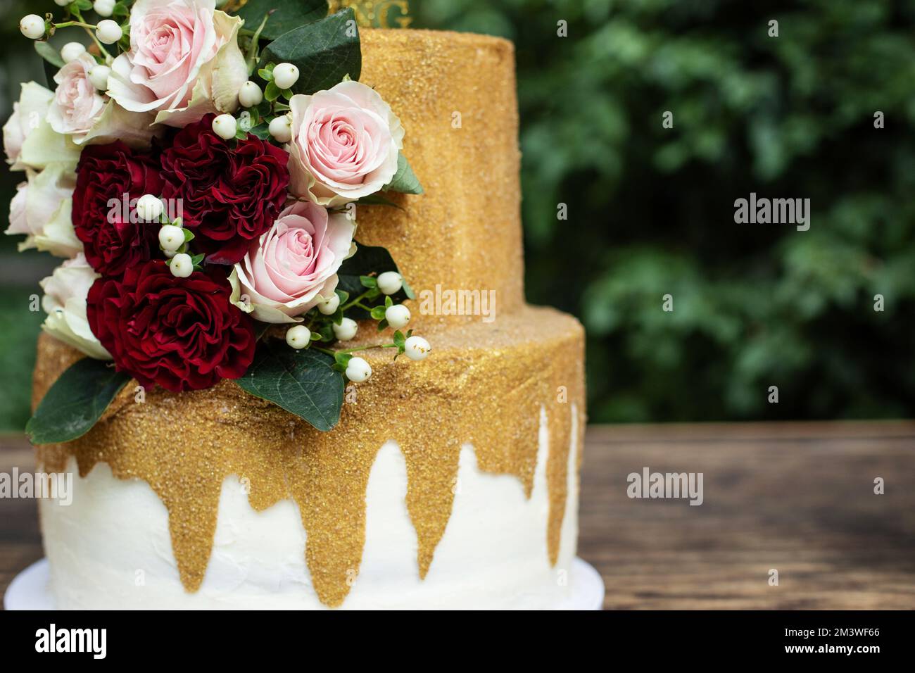 Zweistufige Hochzeitstorte in Weiß und Gold mit roten und pinkfarbenen Rosen und Zruskusblättern Stockfoto