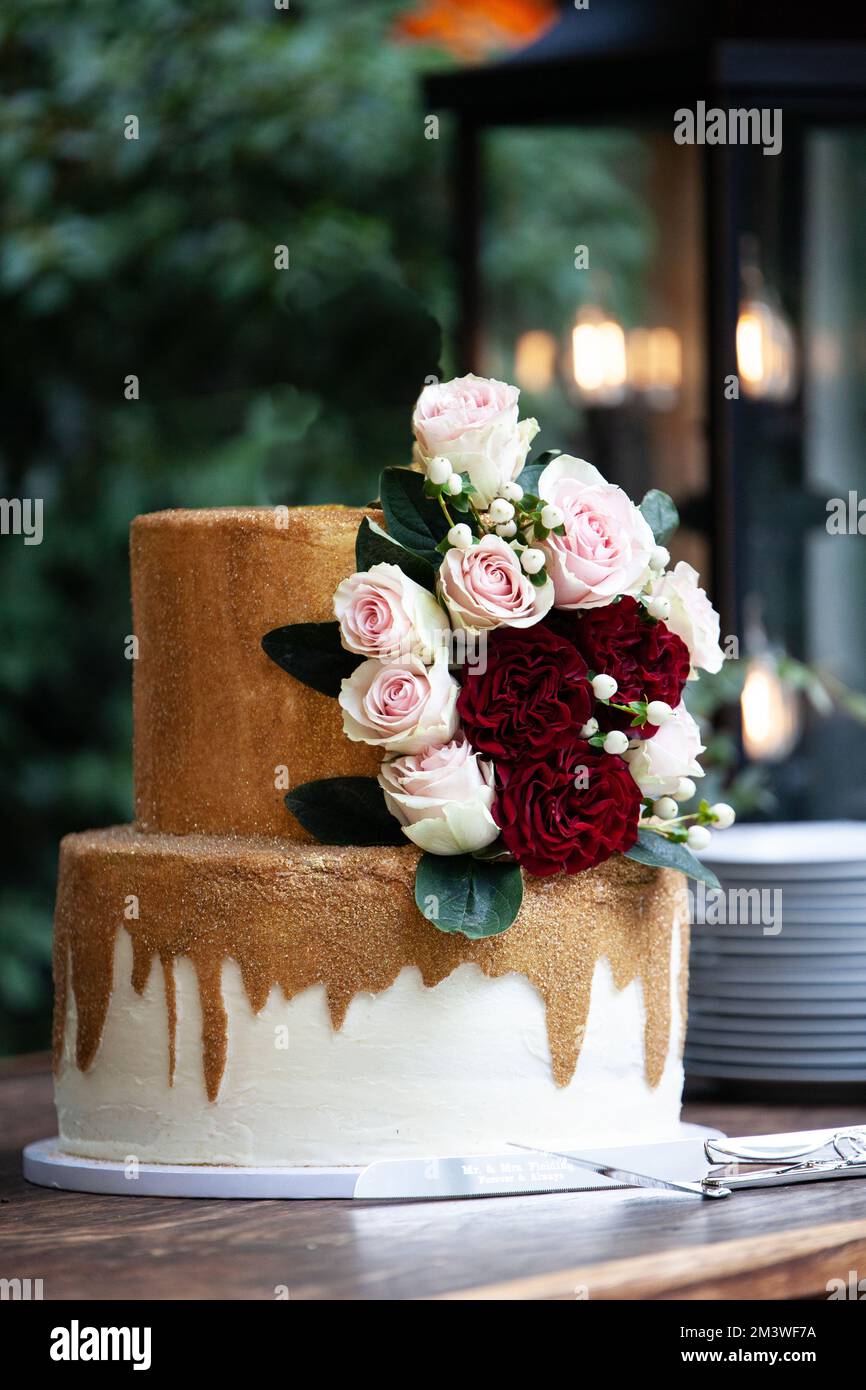 Zweistufige Hochzeitstorte in Weiß und Gold mit roten und pinkfarbenen Rosen und Zruskusblättern Stockfoto