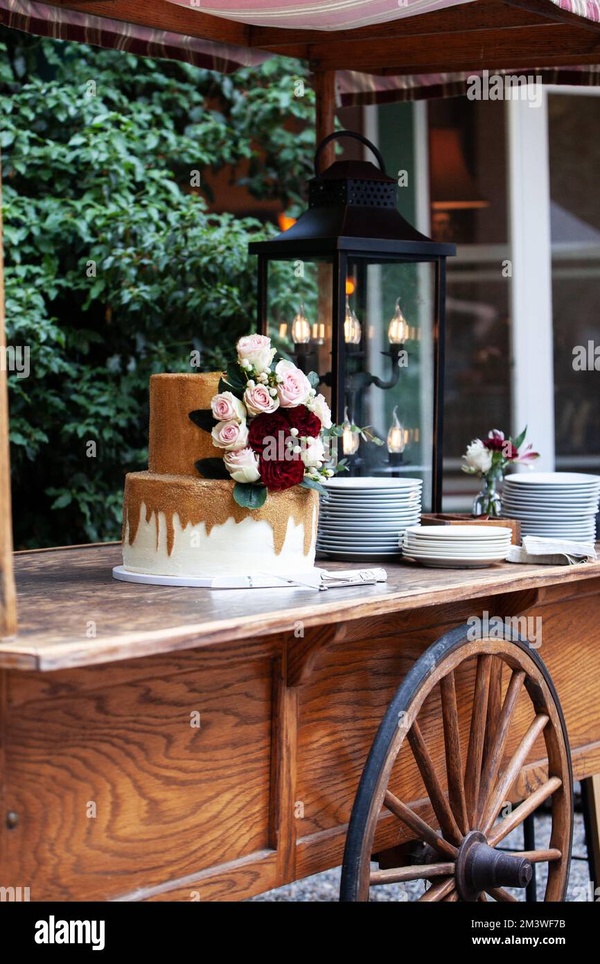Zweistufige Hochzeitstorte in Weiß und Gold mit roten und pinkfarbenen Rosen und Zruskusblättern auf einem Holzwagen Stockfoto