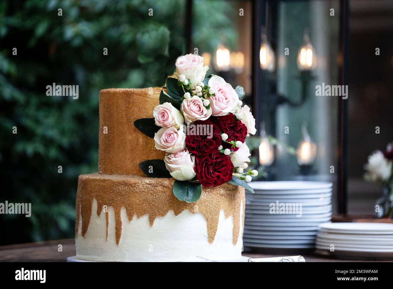 Zweistufige Hochzeitstorte in Weiß und Gold mit roten und pinkfarbenen Rosen und Zruskusblättern auf einem Holzwagen Stockfoto