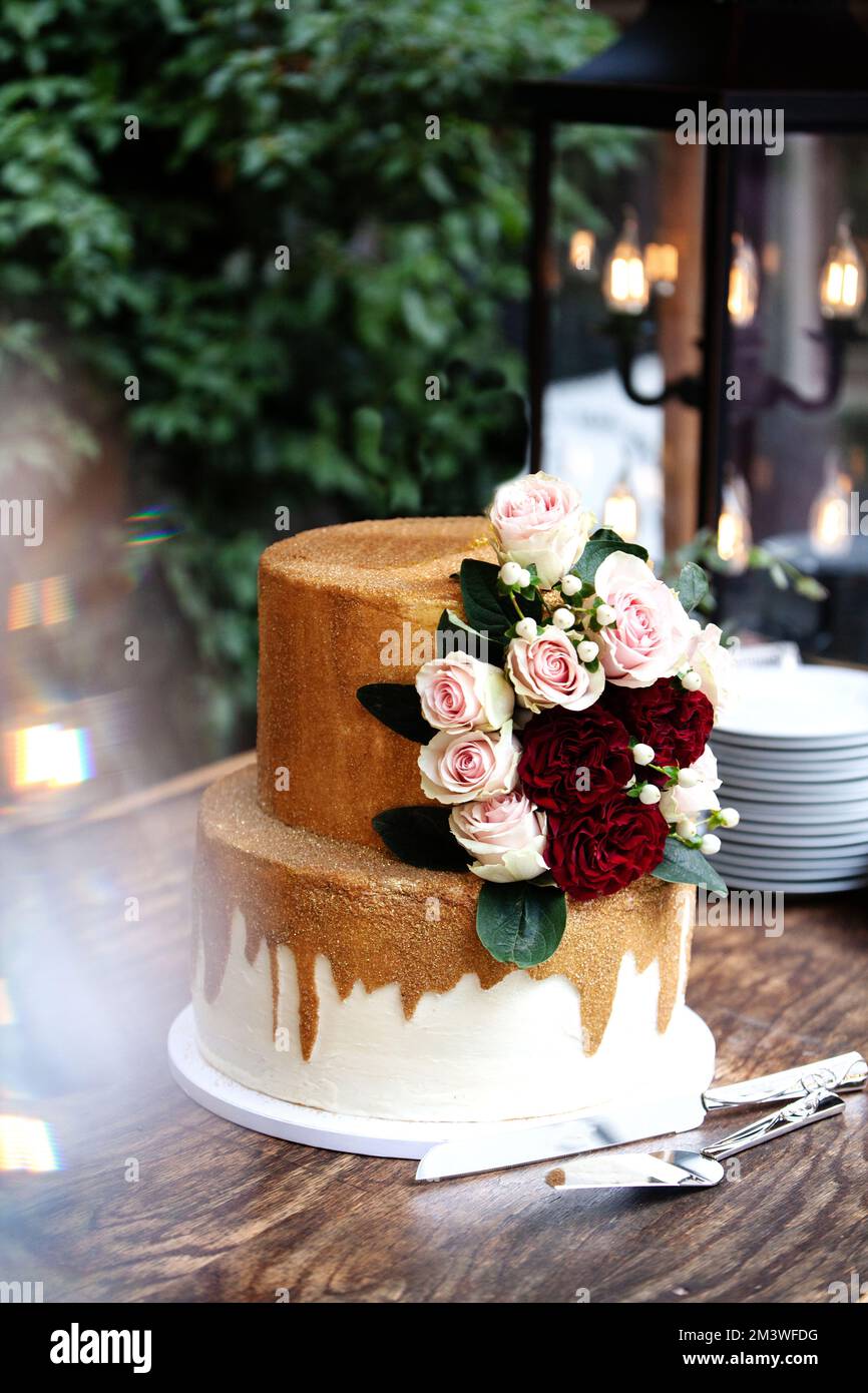 Zweistufige Hochzeitstorte in Weiß und Gold mit roten und pinkfarbenen Rosen und Zruskusblättern auf einem Holzwagen Stockfoto