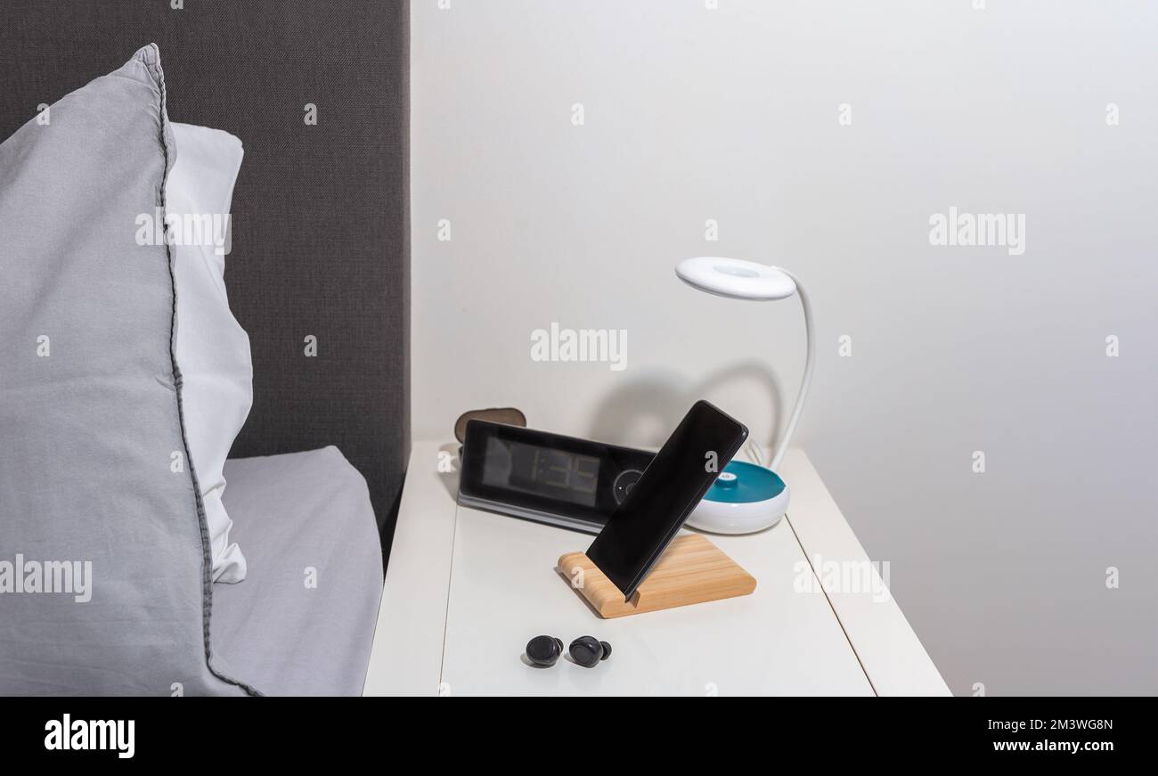 Smartphone auf dem Nachttisch mit Lampe, Headset, Uhr und Bett als Hintergrund Stockfoto