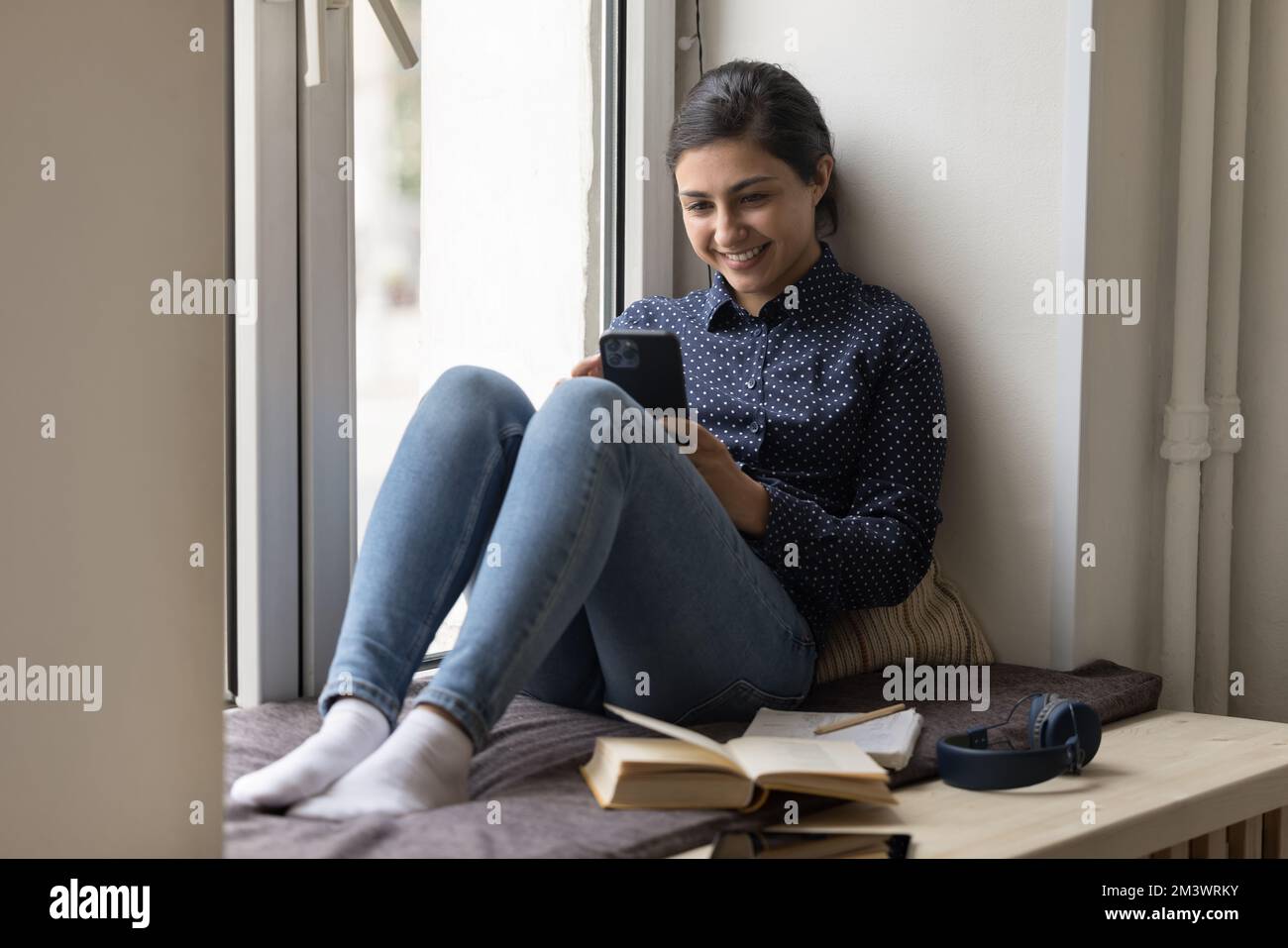 Glückliche Studentin, die sich auf dem Fensterbrett mit dem Handy ausruht Stockfoto