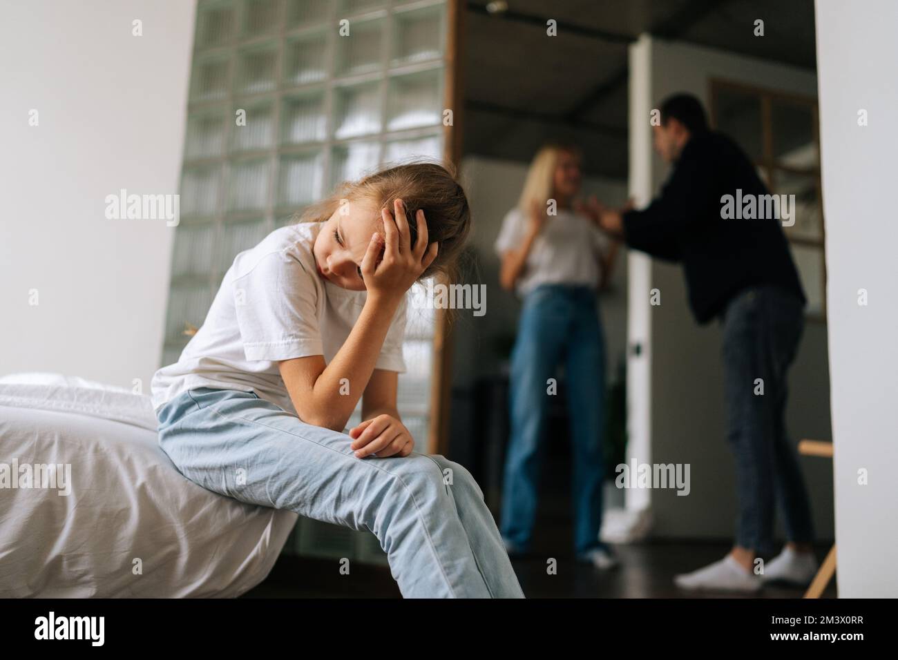 Porträt eines traurigen, einsamen, weinenden kleinen Mädchens, das auf dem Sofa sitzt, während Eltern sich im Wohnzimmer im Hintergrund streiten und streiten. Stockfoto