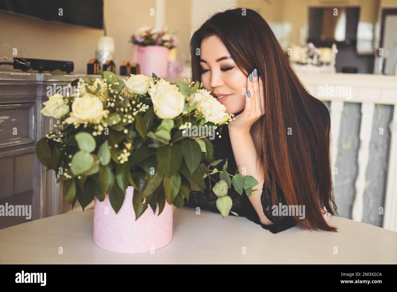 Eine wunderschöne asiatische Frau sitzt am Tisch im Wohnzimmer. Die helle Inneneinrichtung bewundert den Blumenstrauß der weißen Rosen. Lächelnde junge Dame, die ein Überraschungsgeschenk erhalten hat Stockfoto