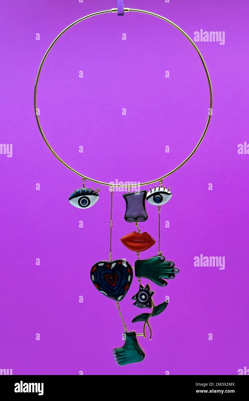 Auffällige runde goldene Halskette mit Augen, Nase, Mund, Herz, Hand, Fuß und Blume. Schmuck mit getrennten Körperteilen vor elektrischem lila Hintergrund. Stockfoto