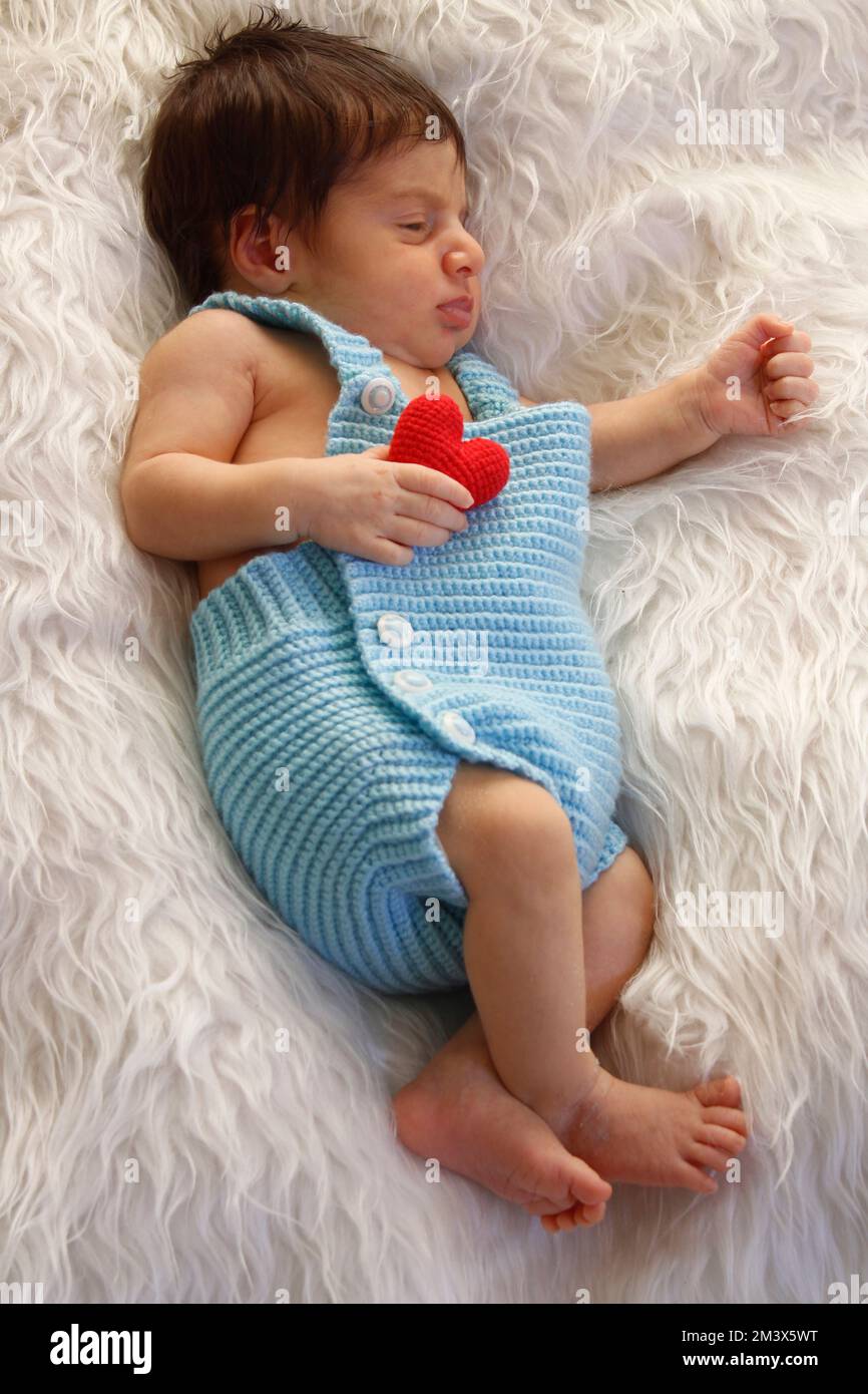 Süßes Baby schläft auf weißer, flauschiger Decke und hält ein kleines rotes Herz in der Hand. Liebe und Gesundheitskonzept. Stockfoto