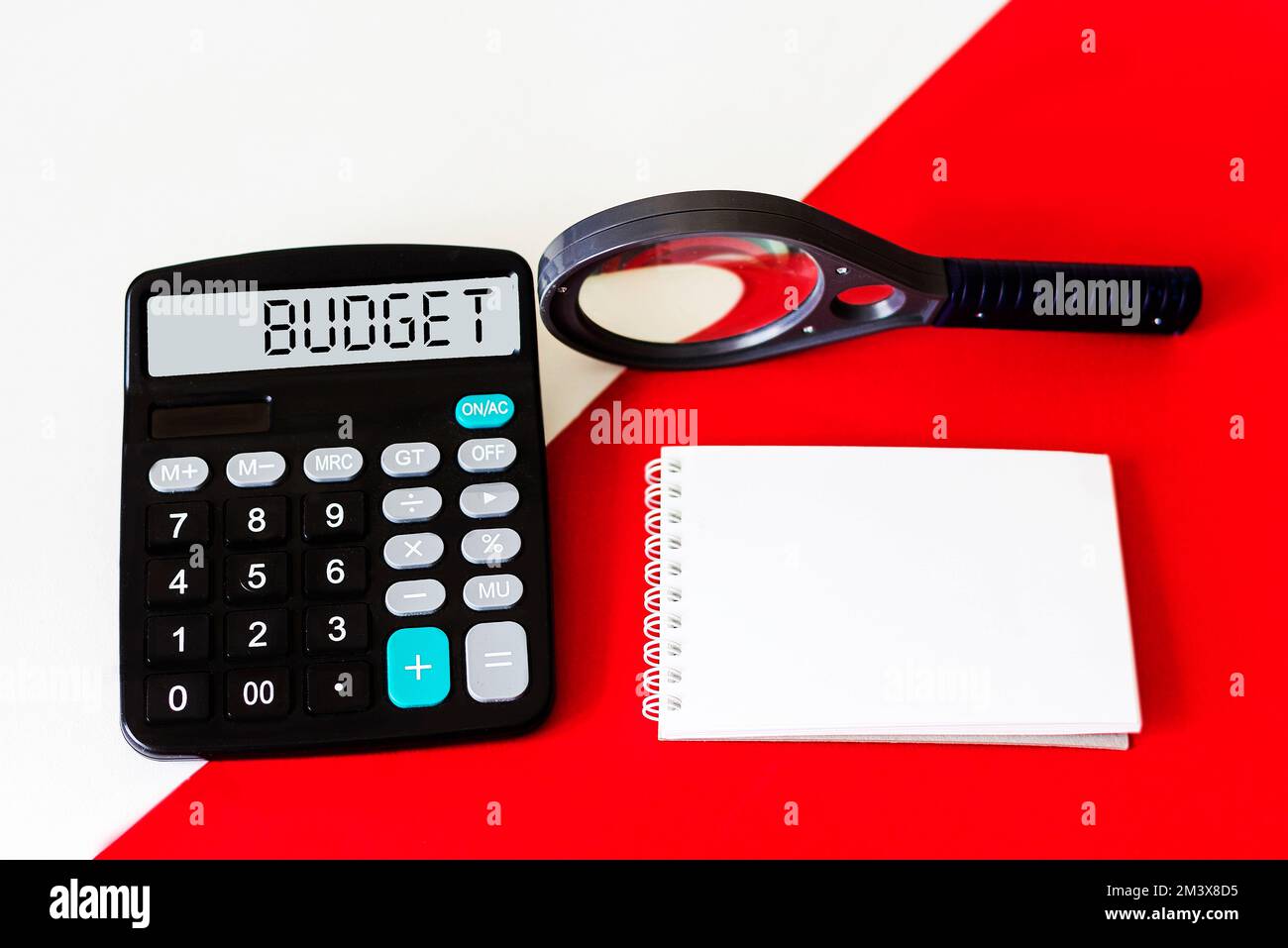 Rechner mit dem Wort BUDGET auf dem Display. Vergrößerungsglas und Notizblock auf rot-weißem Hintergrund. Konzeptfoto Stockfoto