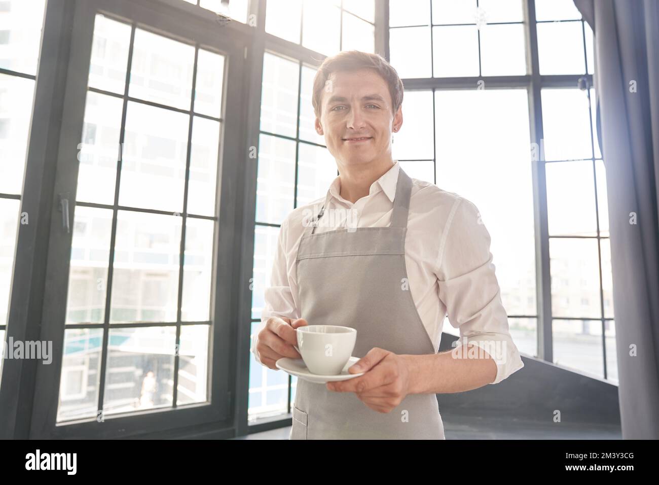 Ein Kellner in einer grauen Schürze hält eine Tasse Kaffee. Ein Barista serviert eine Tasse heißen Kaffee in einem Café vor dem Hintergrund eines großen Fensters. Happy Breakfast Konzept. Hochwertiges Foto Stockfoto