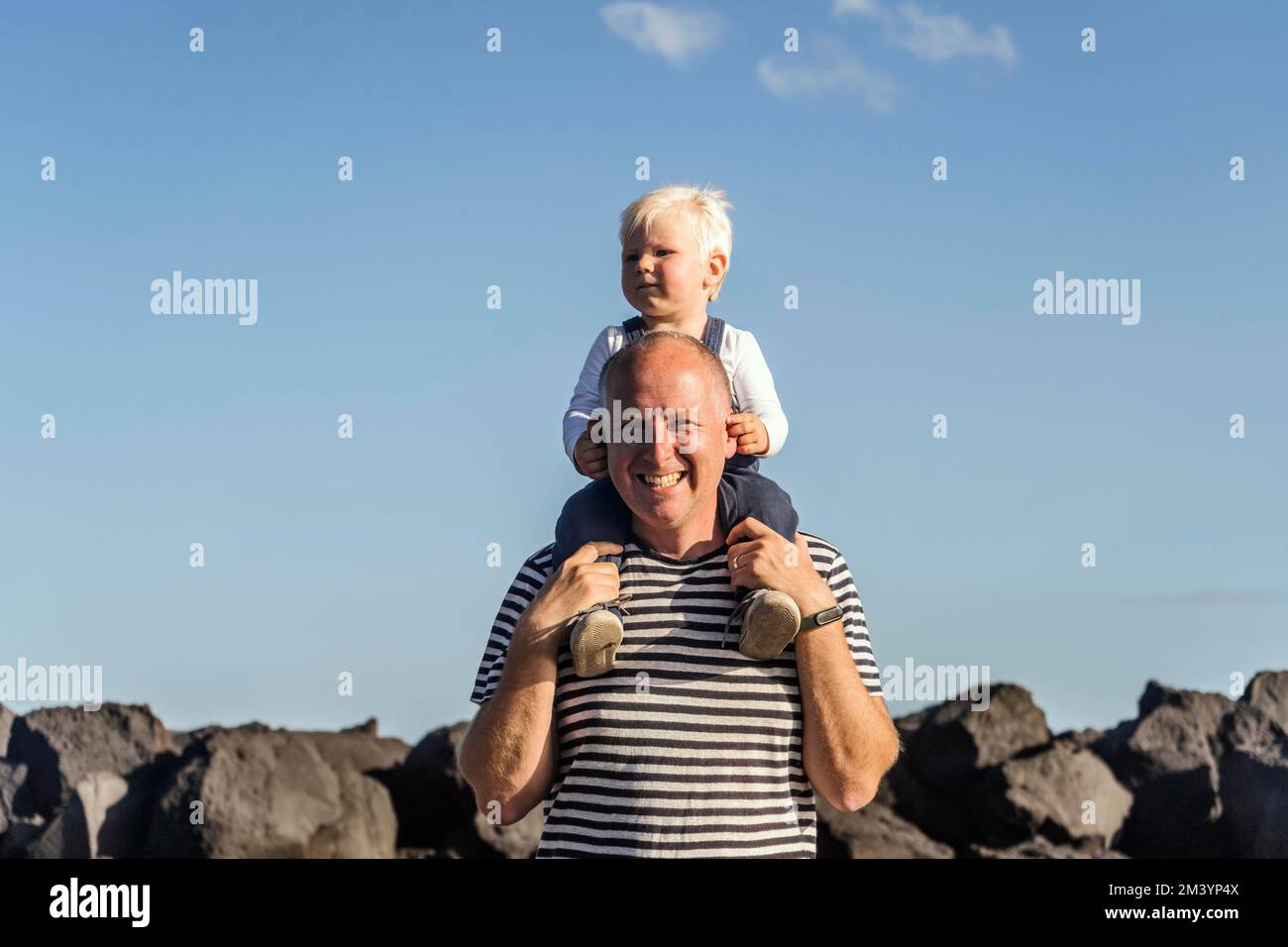 Glücklicher Vater, der seinen kleinen blonden Sohn auf der Schulter gegen den blauen Himmel hält Stockfoto