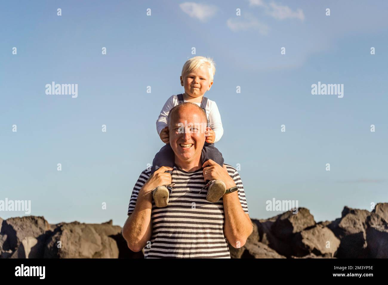Glücklicher Vater, der seinen kleinen blonden Sohn auf der Schulter gegen den blauen Himmel hält Stockfoto