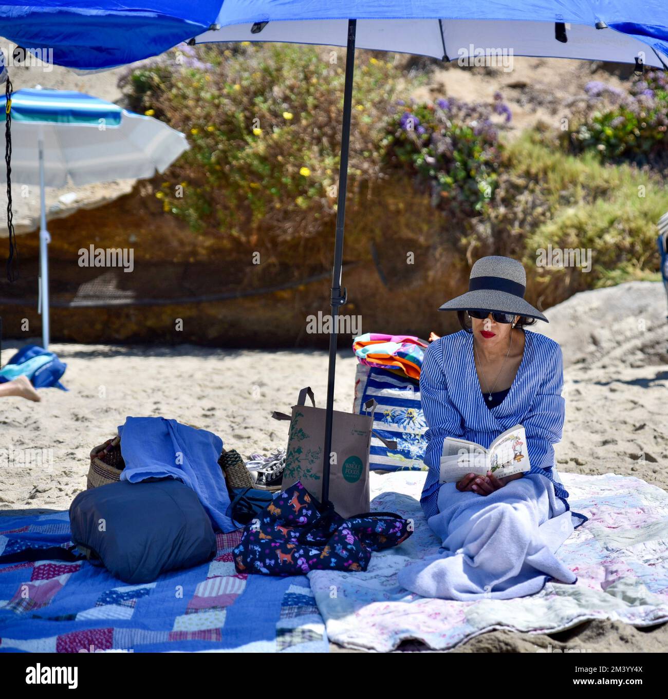 Eine stilvoll gekleidete Erwachsene Frau, die unter einem blauen Sonnenschirm am Strand sitzt und sich beim Lesen eines Buches entspannt Stockfoto