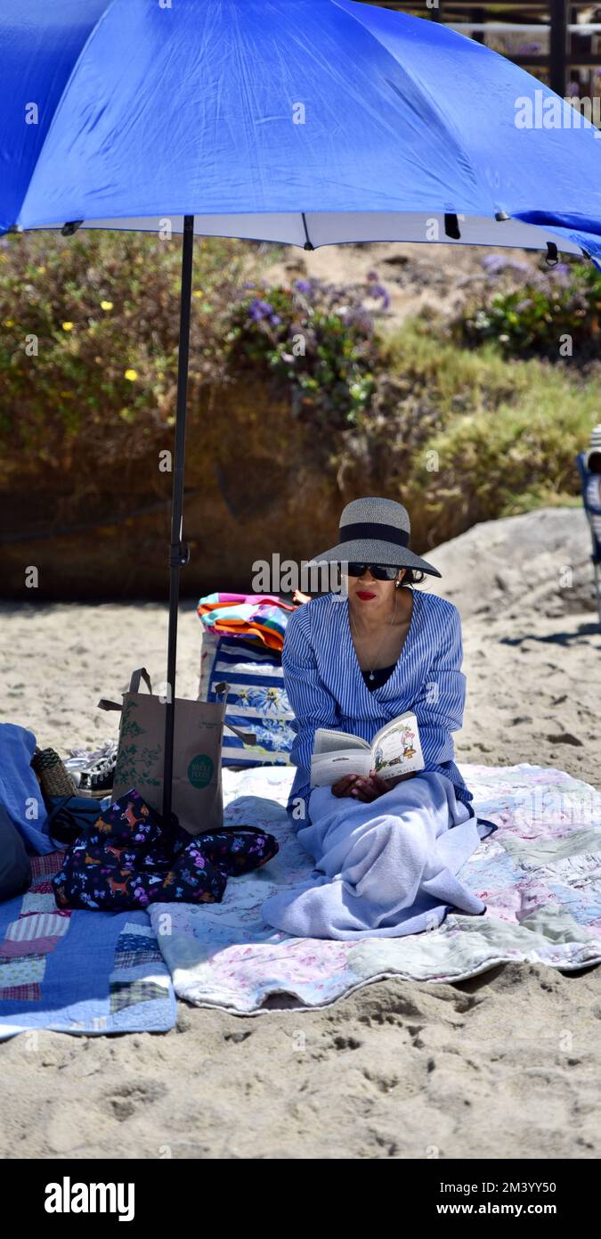 Eine stilvoll gekleidete Erwachsene Frau, die unter einem blauen Sonnenschirm am Strand sitzt und sich beim Lesen eines Buches entspannt Stockfoto