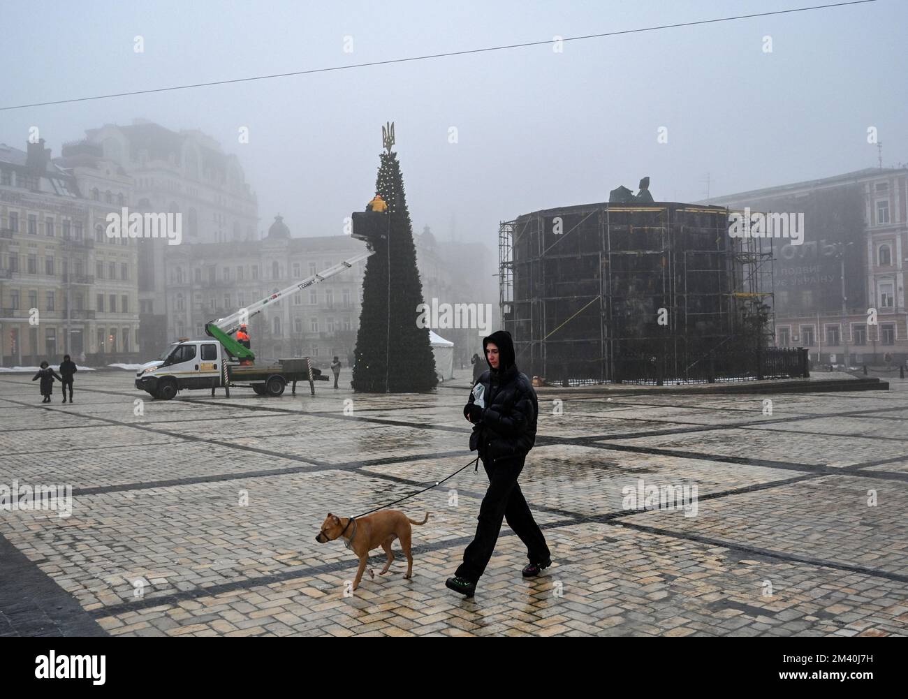 Kiew, Ukraine. 17.. Dezember 2022. Eine Frau mit einem Hund geht an einem Weihnachtsbaum vorbei, der von Stadtarbeitern dekoriert wird, in der Nähe eines Denkmals für Bohdan Chmelnytsky, geschützt durch eine Schutzstruktur vor Raketenangriffen der russischen Armee während des Stromausfalls in Kiew. Die russische Armee führte massive Raketenangriffe auf die ukrainische Energieinfrastruktur durch. Nach schweren Schäden am Stromnetz in vielen Städten der Ukraine führte die National Power Company Ukrenergo Notfall- und stündliche Stromausfälle ein. (Foto: Sergei Chuzavkov/SOPA Images/Sipa USA) Guthaben: SIPA USA/Alamy Live News Stockfoto