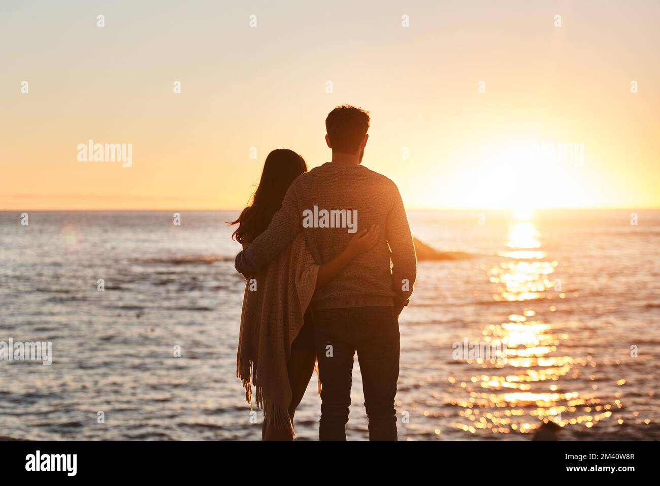 Nichts inspiriert Romantik so wie ein Sonnenuntergang. Rückblick auf ein glückliches junges Paar, das den Sonnenuntergang am Strand beobachtet. Stockfoto