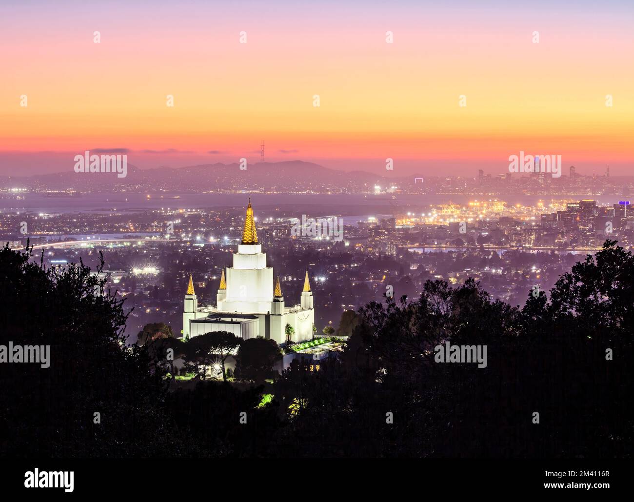 Der Oakland California Temple in den Hügeln von Oakland und die Skyline der Stadt unter farbenfrohem Himmel in der Abenddämmerung, Kalifornien Stockfoto