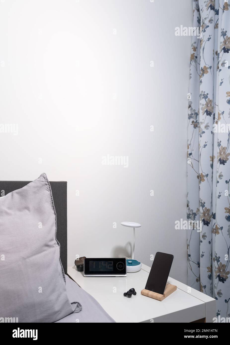 Smartphone auf dem Nachttisch mit Lampe, Headset, Uhr und Bett als Hintergrund. Vertikal Stockfoto