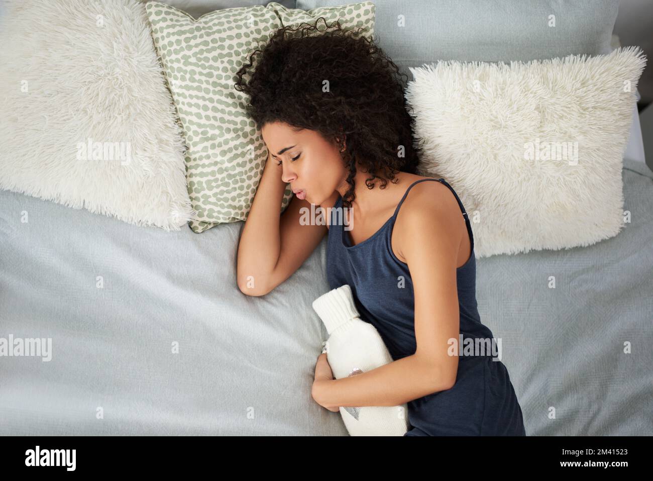 Durch den Schmerz atmen. Eine attraktive junge Frau, die unter Periodenkrämpfen leidet, während sie mit einer Wärmflasche im Bett liegt. Stockfoto