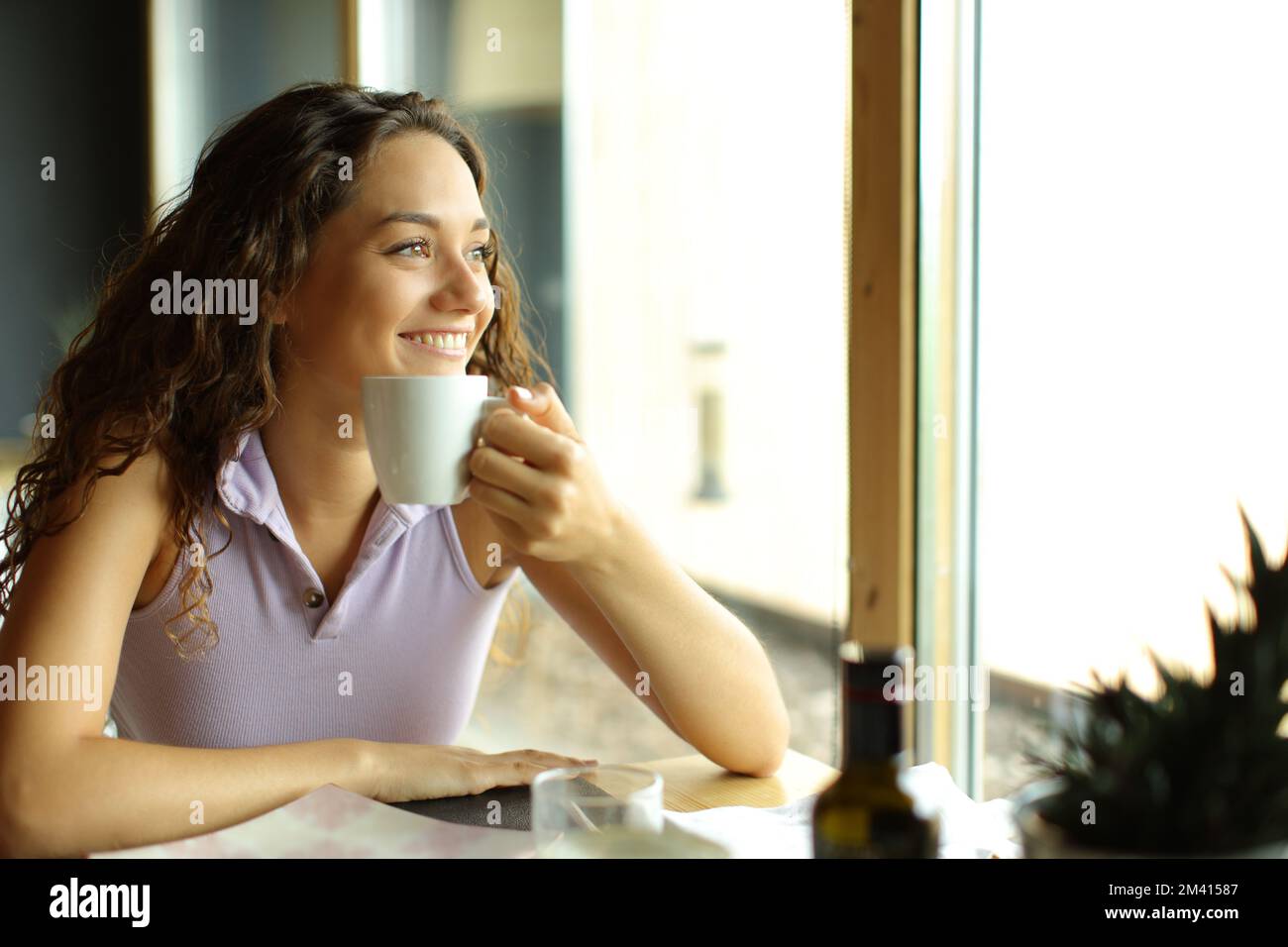 Eine glückliche Frau, die eine Kaffeetasse in einem Restaurant hält, blickt durch ein Fenster weg Stockfoto