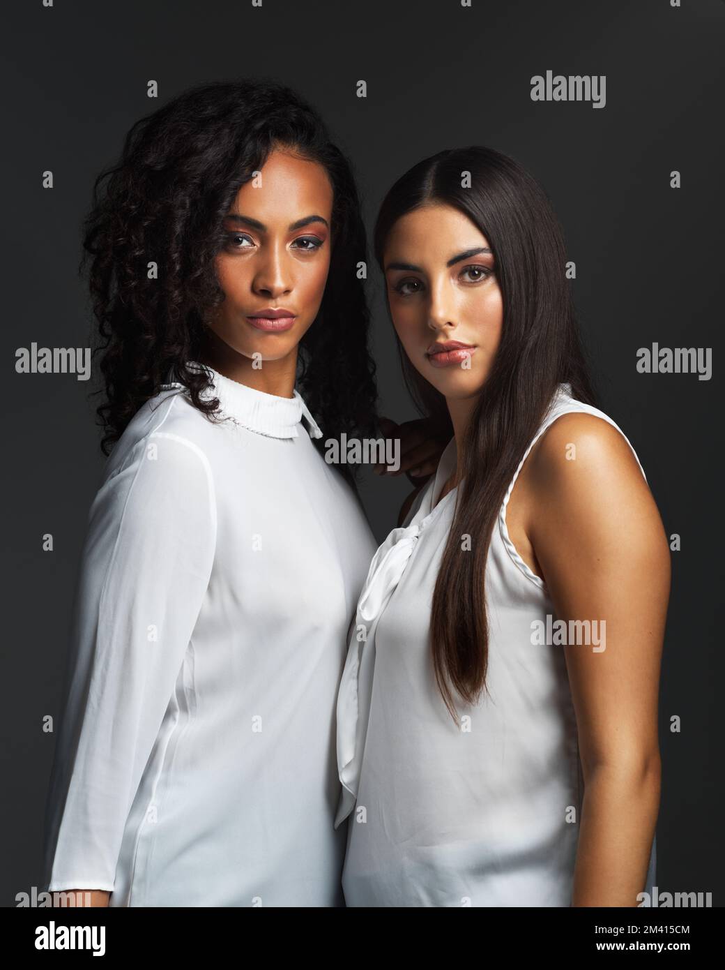 Zusammenstehen und aufrecht stehen. Porträt von zwei attraktiven jungen Frauen, die weiße Blusen tragen und sich eng beieinander vor einem dunklen Hintergrund in posten Stockfoto