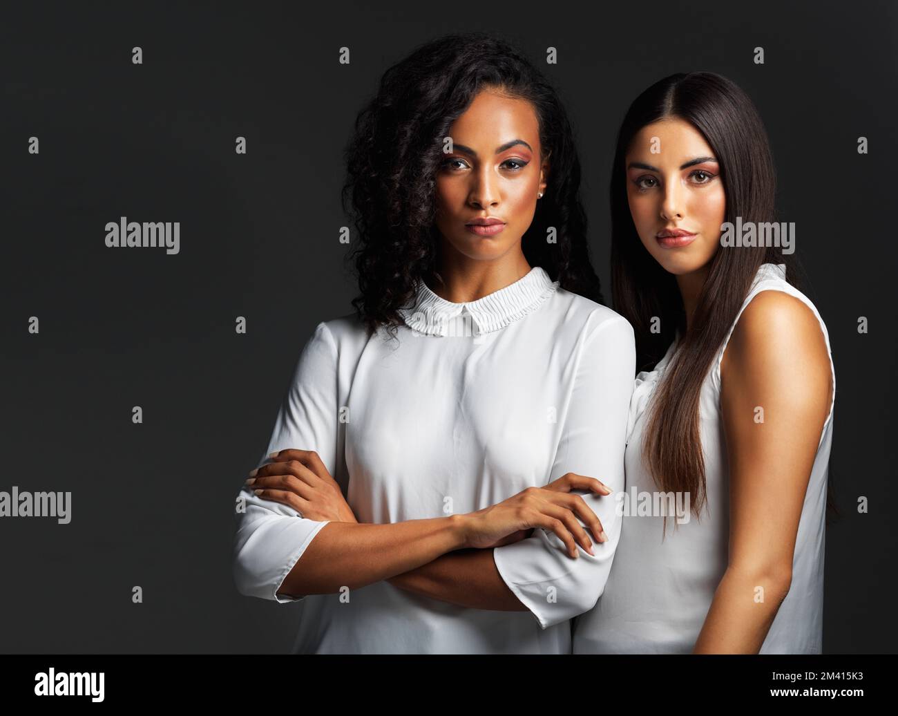 Wir sind schön, selbstbewusst und können nicht aufgehalten werden. Porträt von zwei attraktiven jungen Frauen, die weiße Blusen tragen und sich eng gegen eine Dunkelheit postieren Stockfoto