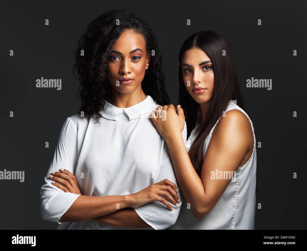 Sie haben das Selbstvertrauen, ihrer Schönheit zu folgen. Porträt von zwei attraktiven jungen Frauen, die weiße Blusen tragen und sich eng gegen eine Dunkelheit postieren Stockfoto