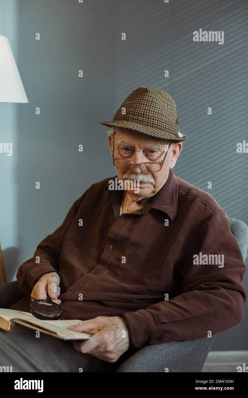 Ein älterer Mann, der allein auf einem Sessel im Wohnzimmer sitzt, eine Brille trägt und ein Buch hält, mit Bedacht in die Kamera schaut Stockfoto