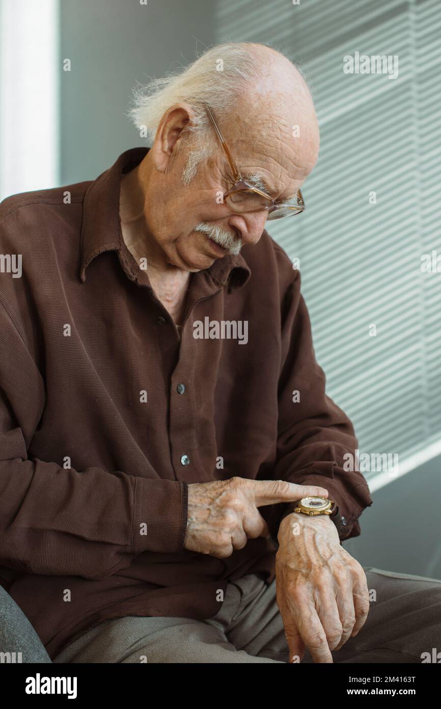 Ein einsamer alter Mann im Wohnzimmer sieht auf seine Uhr am Handgelenk. Warten auf den Besuch eines Freundes oder Verwandter. Stockfoto