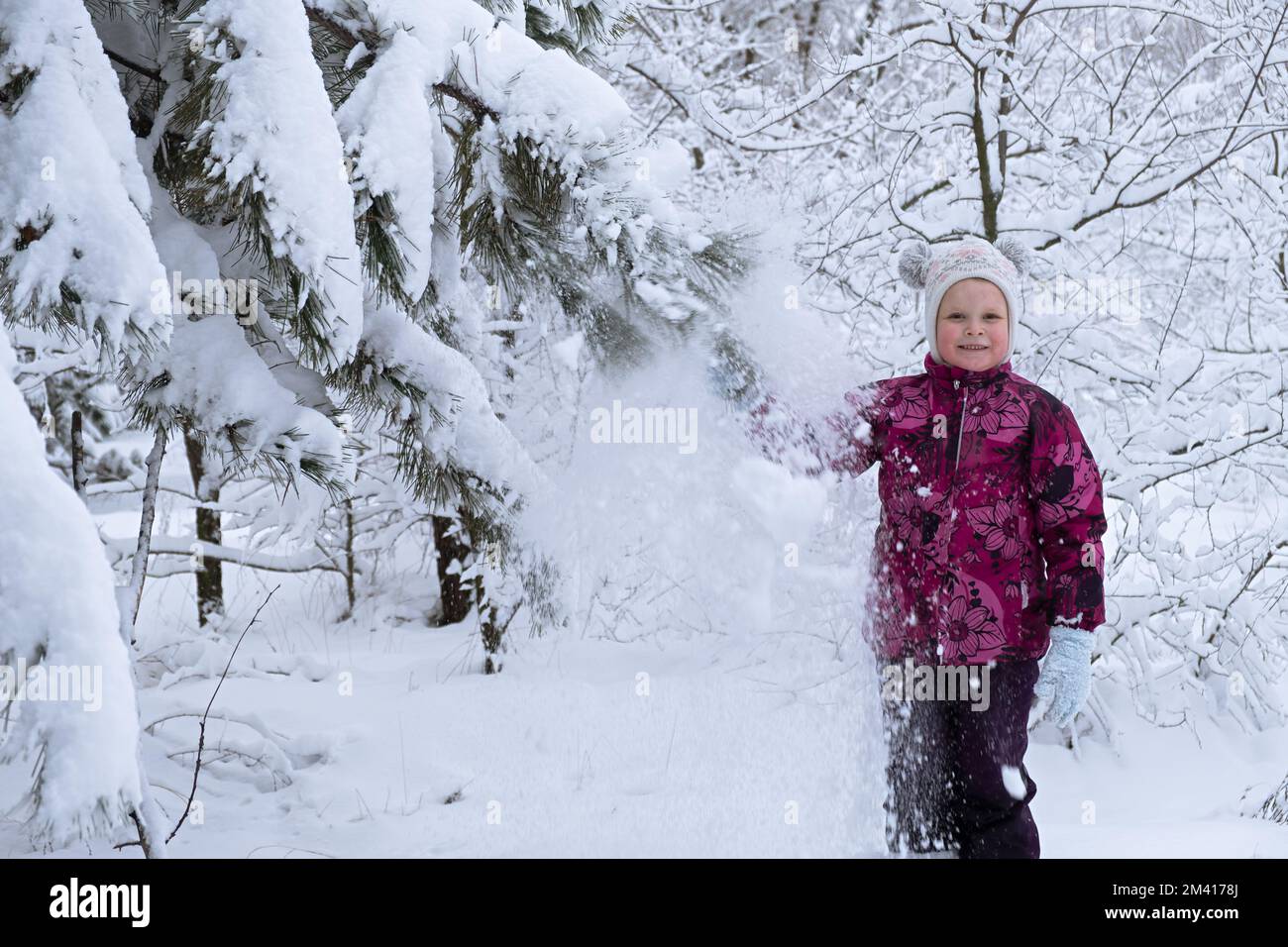 Kinderschnee macht Spaß. Ein kleines Mädchen in einem Winterwald. Er wirft Schnee von den Tannenästen und lacht. Das Konzept der Kindheit, Nachlässigkeit und Stockfoto