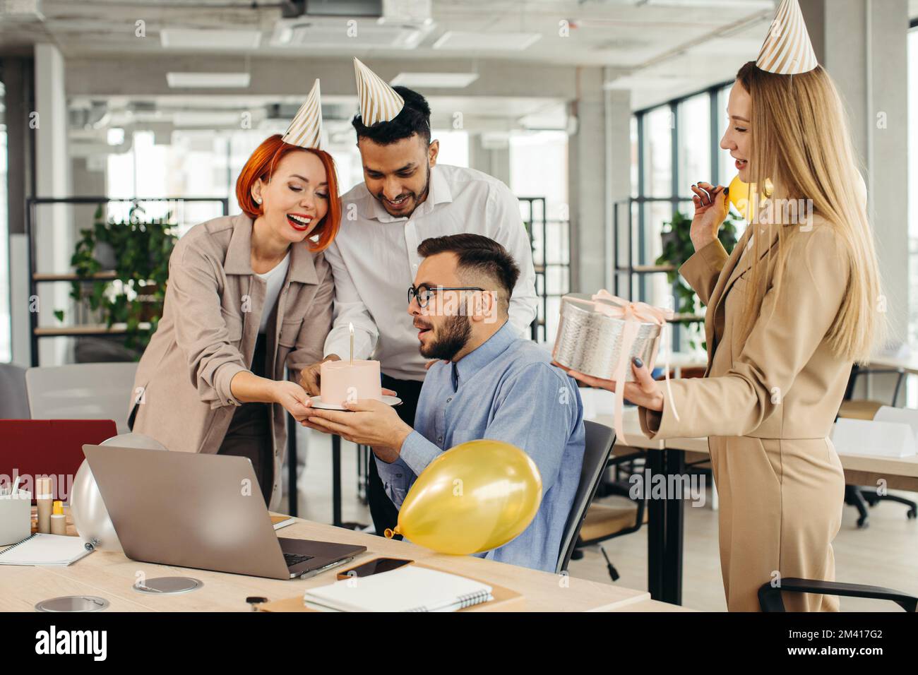 Ein glücklicher Geschäftsmann bläst eine Kerze auf dem Kuchen aus, während er mit weiblichen Kollegen im Büro Geburtstag feiert. Stockfoto