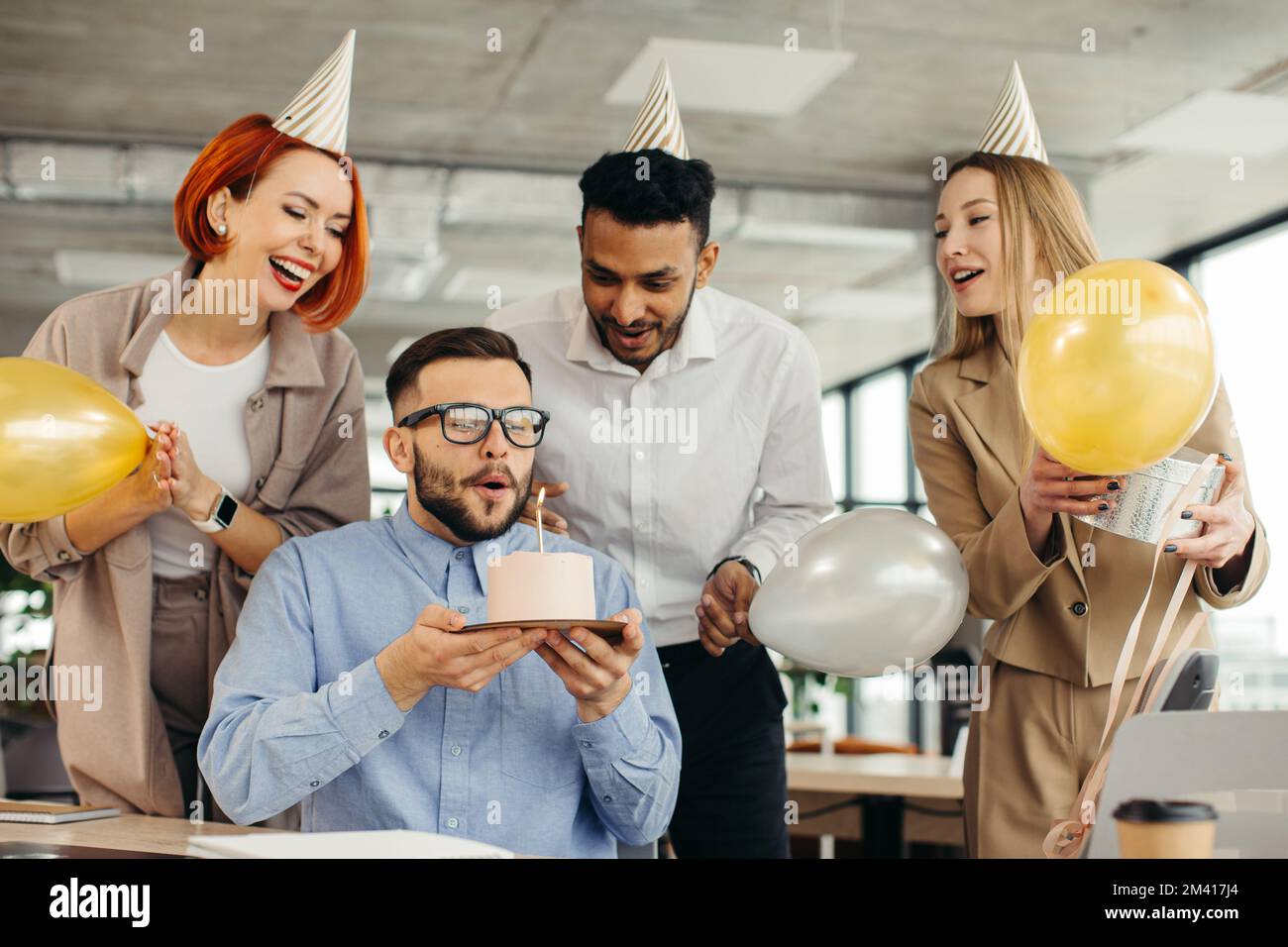 Ein glücklicher Geschäftsmann bläst eine Kerze auf dem Kuchen aus, während er mit weiblichen Kollegen im Büro Geburtstag feiert. Stockfoto