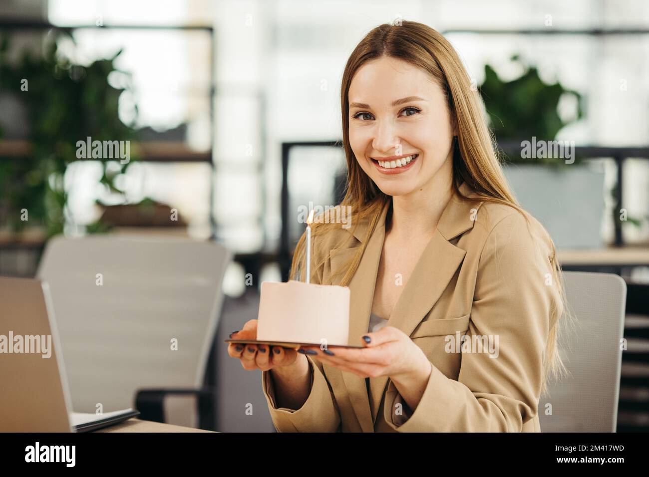 Glückliche Geschäftsfrau, die im Büro mit einer Kerze Kuchen hält und in die Kamera schaut. Stockfoto