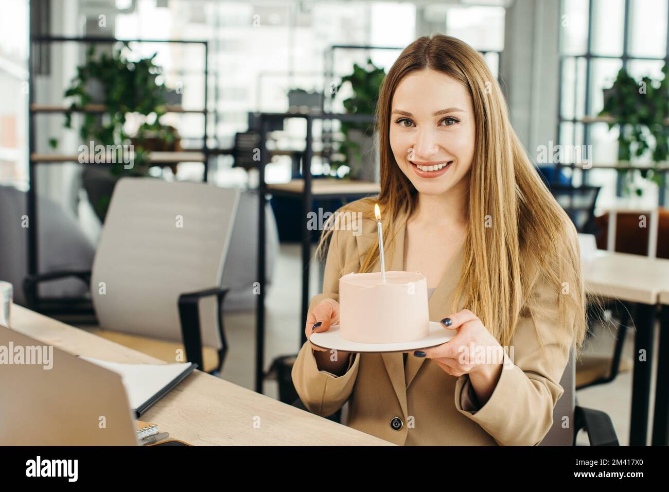 Glückliche Geschäftsfrau, die im Büro mit einer Kerze Kuchen hält und in die Kamera schaut. Stockfoto