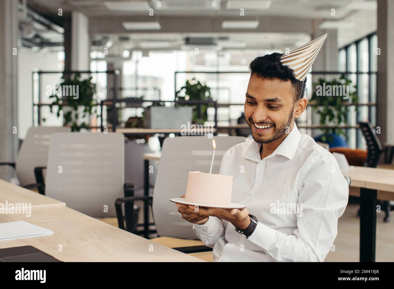 Ein glücklicher hinduistischer Geschäftsmann, der im Büro mit einer Kerze Kuchen hält und in die Kamera schaut. Stockfoto