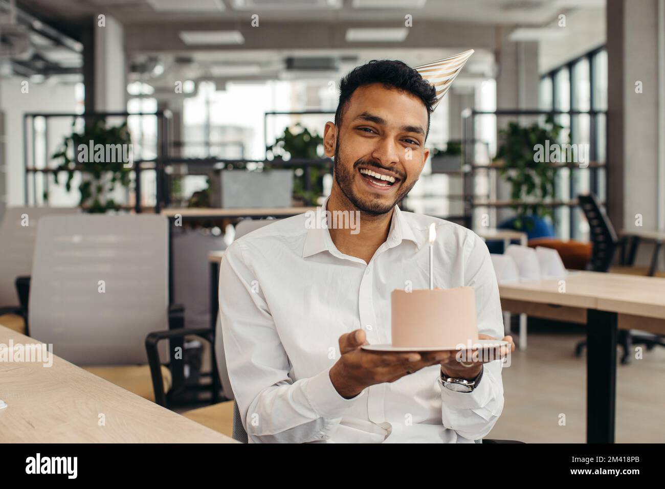 Ein glücklicher hinduistischer Geschäftsmann, der im Büro mit einer Kerze Kuchen hält und in die Kamera schaut. Stockfoto