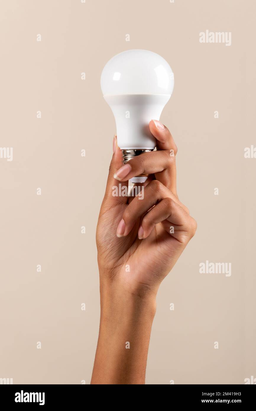 Anonyme schwarze Frau, die LED-Glühbirne in erhobenen Armen vorführt und gleichzeitig eine energiesparende Umweltkampagne vor beigefarbenem Hintergrund fördert Stockfoto