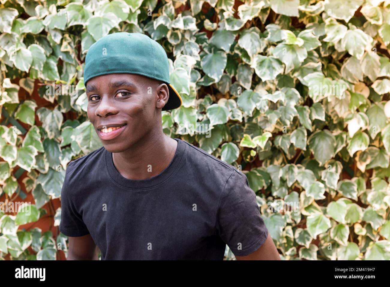 Ein glücklicher afroamerikanischer Mann in T-Shirt und Mütze, der in die Kamera schaut, mit einem Lächeln neben grünen Blättern im Garten Stockfoto