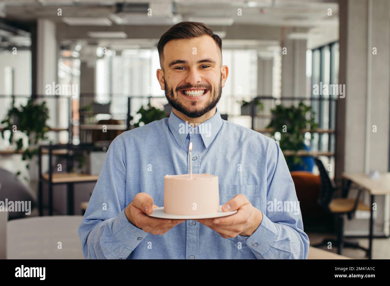 Aufgeregter Mann feiert Geburtstag im Büro, sieht glücklich aus, hält Kuchen mit Kerze in der Hand, wünscht sich was. Stockfoto