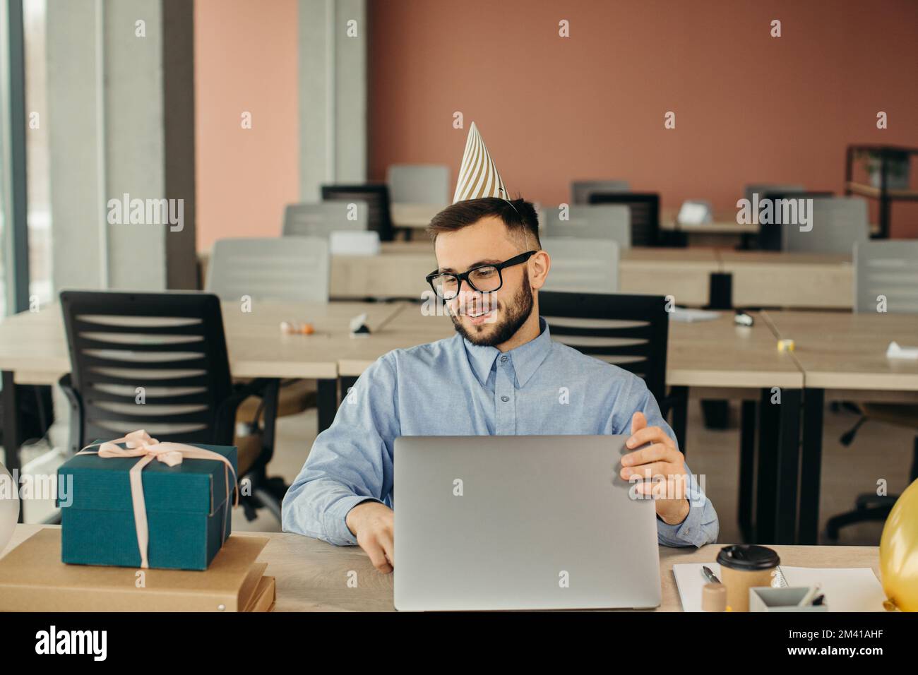 Nach der Geburtstagsparty. Büroangestellter mit Bart und Hut sitzt am Arbeitsplatz mit Laptop und Geschenk mit festlichem Band. Stockfoto