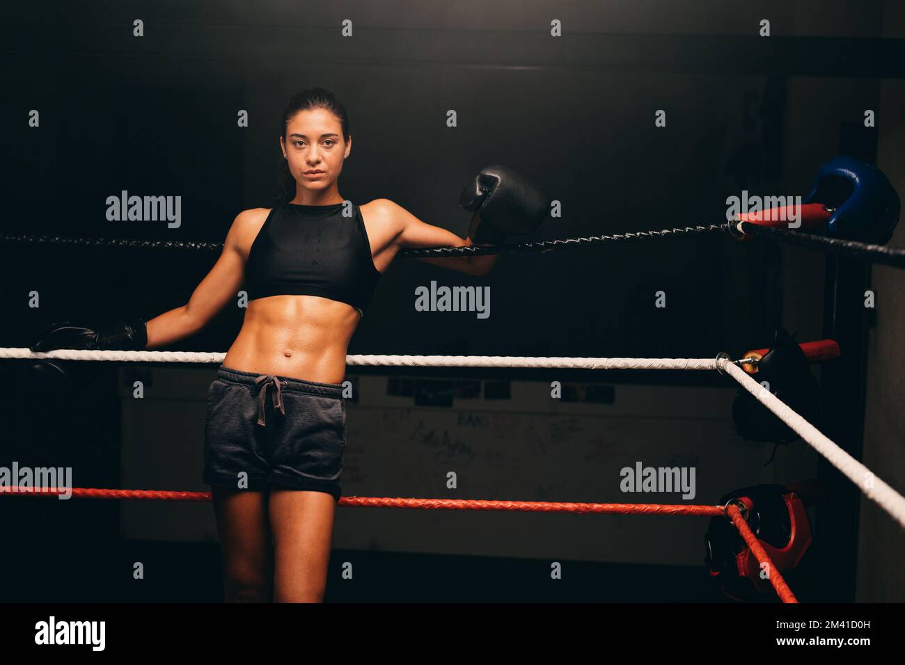 Sportliche junge Frau, die in einem Boxring in die Kamera schaut, während sie an den Seilen steht. Boxertraining in einem Fitnessraum. Stockfoto