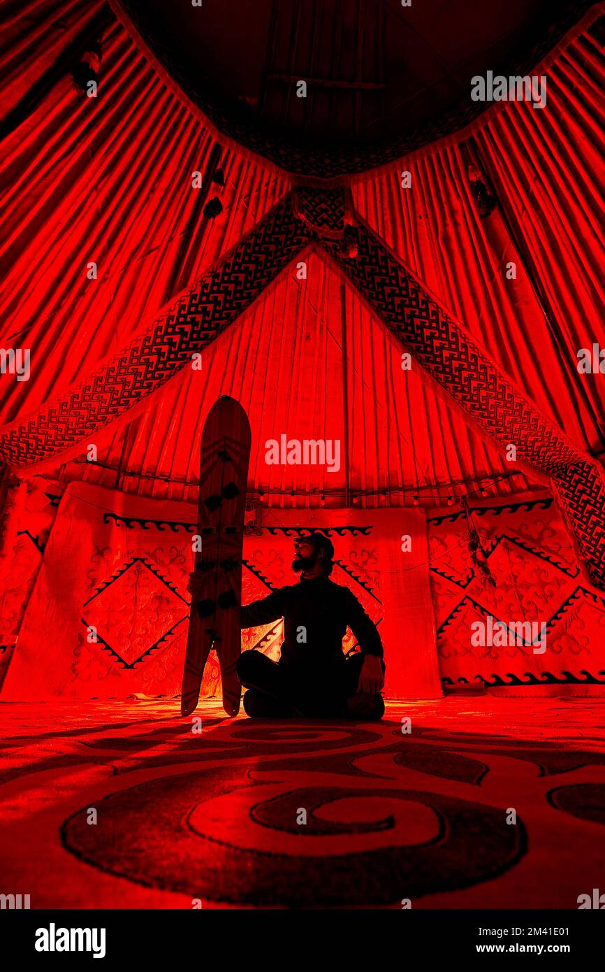 Silhouette von Mann Hold mit geteiltem Snowboard, das mit gekreuzten Beinen im Nomadenhaus Yurt gegen Teppich mit ethnischen Mustern sitzt, die durch rotes Licht beleuchtet werden Stockfoto