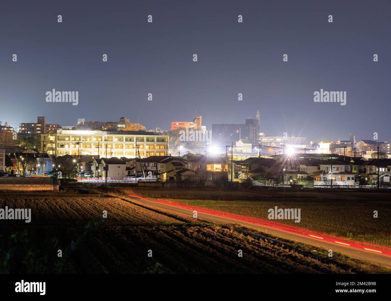 Rote Fahrzeuglichter Streifen nachts durch gepflügte Felder am Stadtrand Stockfoto