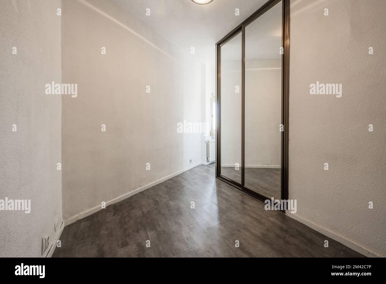 Ein leeres Zimmer mit eingebautem Kleiderschrank mit Spiegelschiebetüren mit ockerfarbenen Ecken aus eloxiertem Aluminium, weiß bemalten Wänden mit rauem Görtchen und grauem w Stockfoto