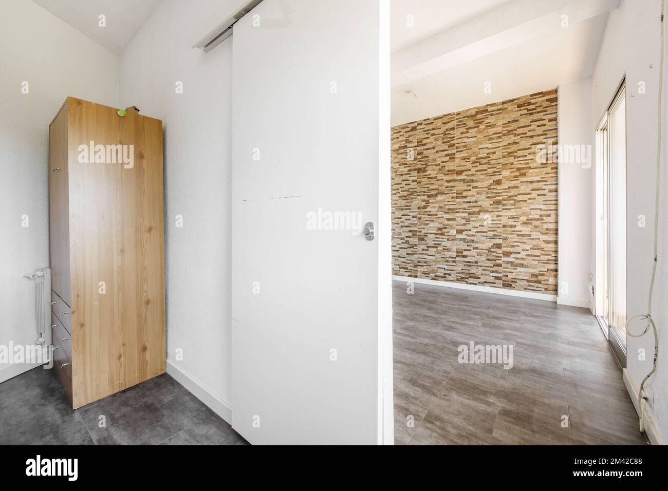Ein leeres Zimmer mit einem Holzschrank in einer Ecke, eine weiße Holzschiebetür, die zu einem anderen Raum mit einer gefliesten Wand ähnlich wie Steinascher führt. Stockfoto