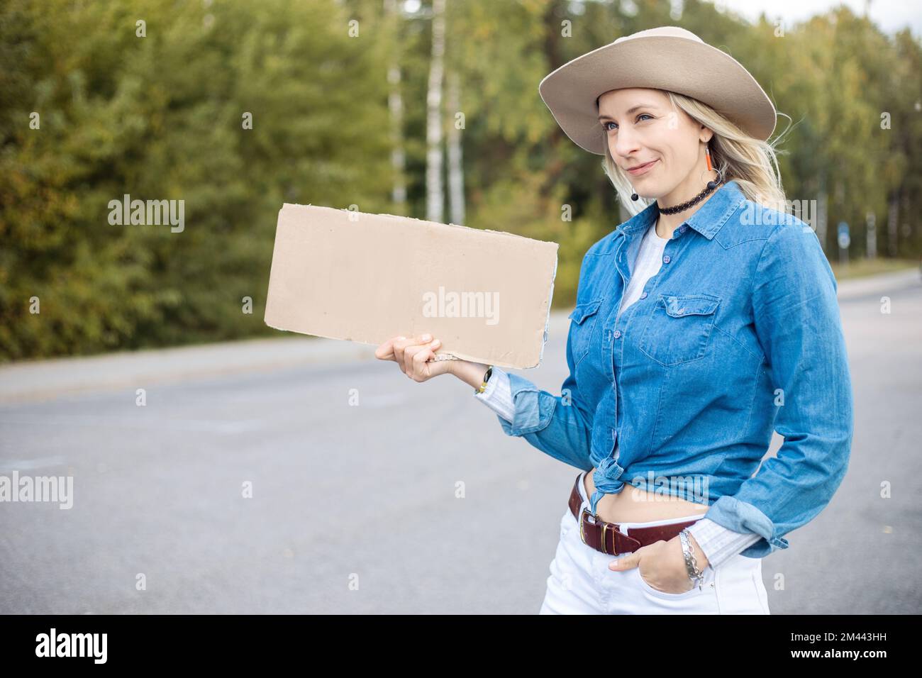 Moderne, sarkastische, blonde Frau mit Hut trampen mit leerem Karton und warten auf Auto-Stopp. Hitchhiker-Risiko Stockfoto
